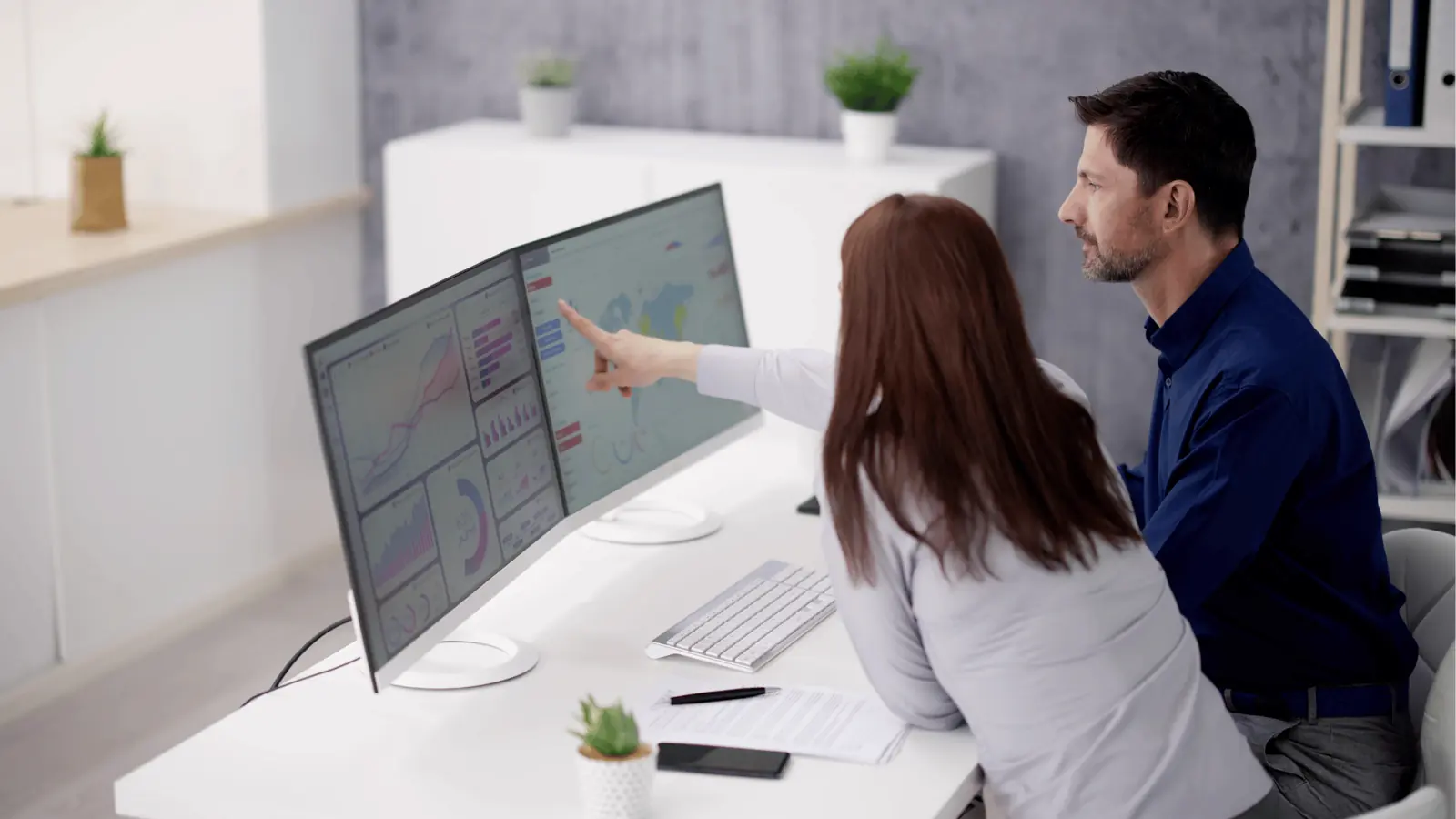 Two colleagues pointing at dual monitors showing charts and maps in office for team collaboration