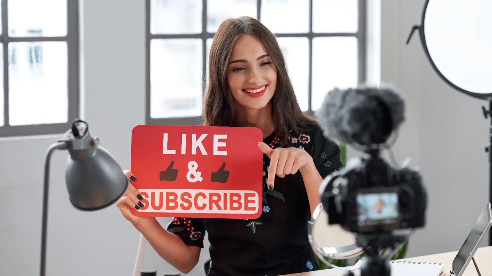 Smiling female content creator holding 'Like & Subscribe' sign in front of camera for publishing