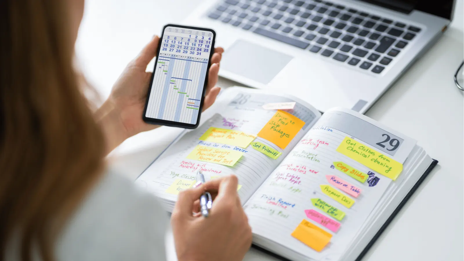 Person holding smartphone showing calendar while writing in sticky-note planner for content calendar