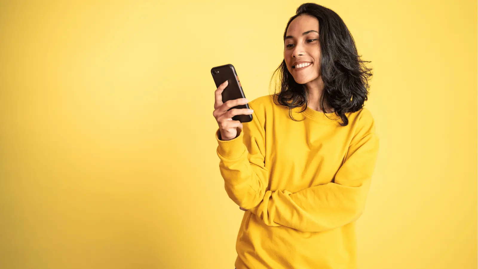 Smiling woman in yellow sweatshirt looking at smartphone against yellow background