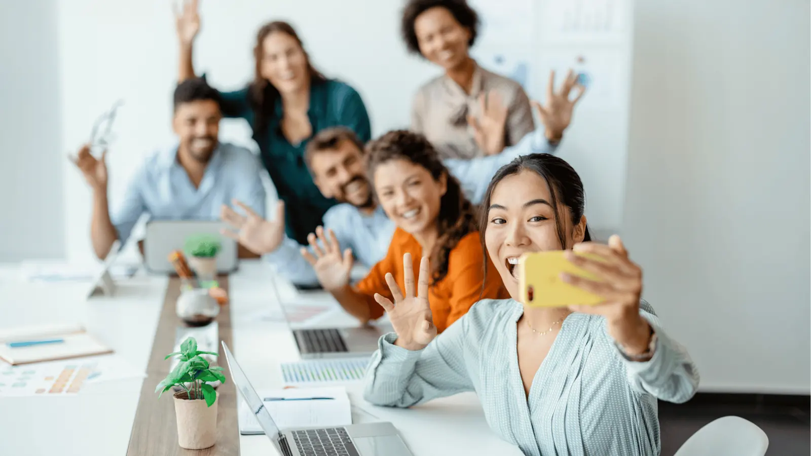 Diverse coworkers smiling and waving while taking a group selfie at desk