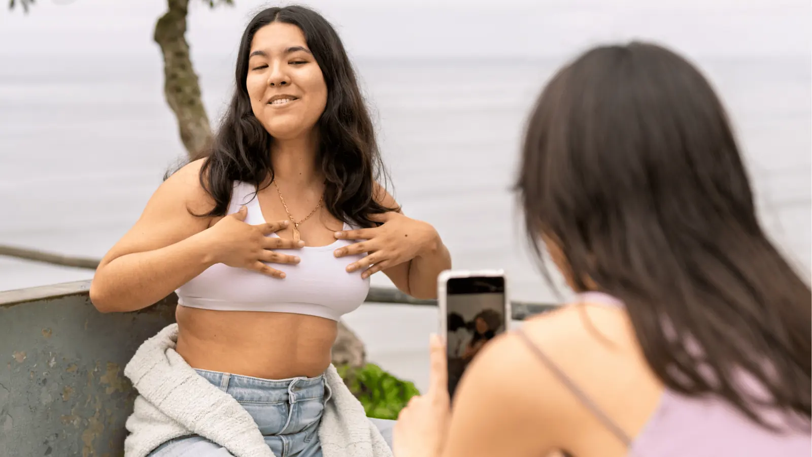 Young woman posing in white crop top while friend photographs her with smartphone