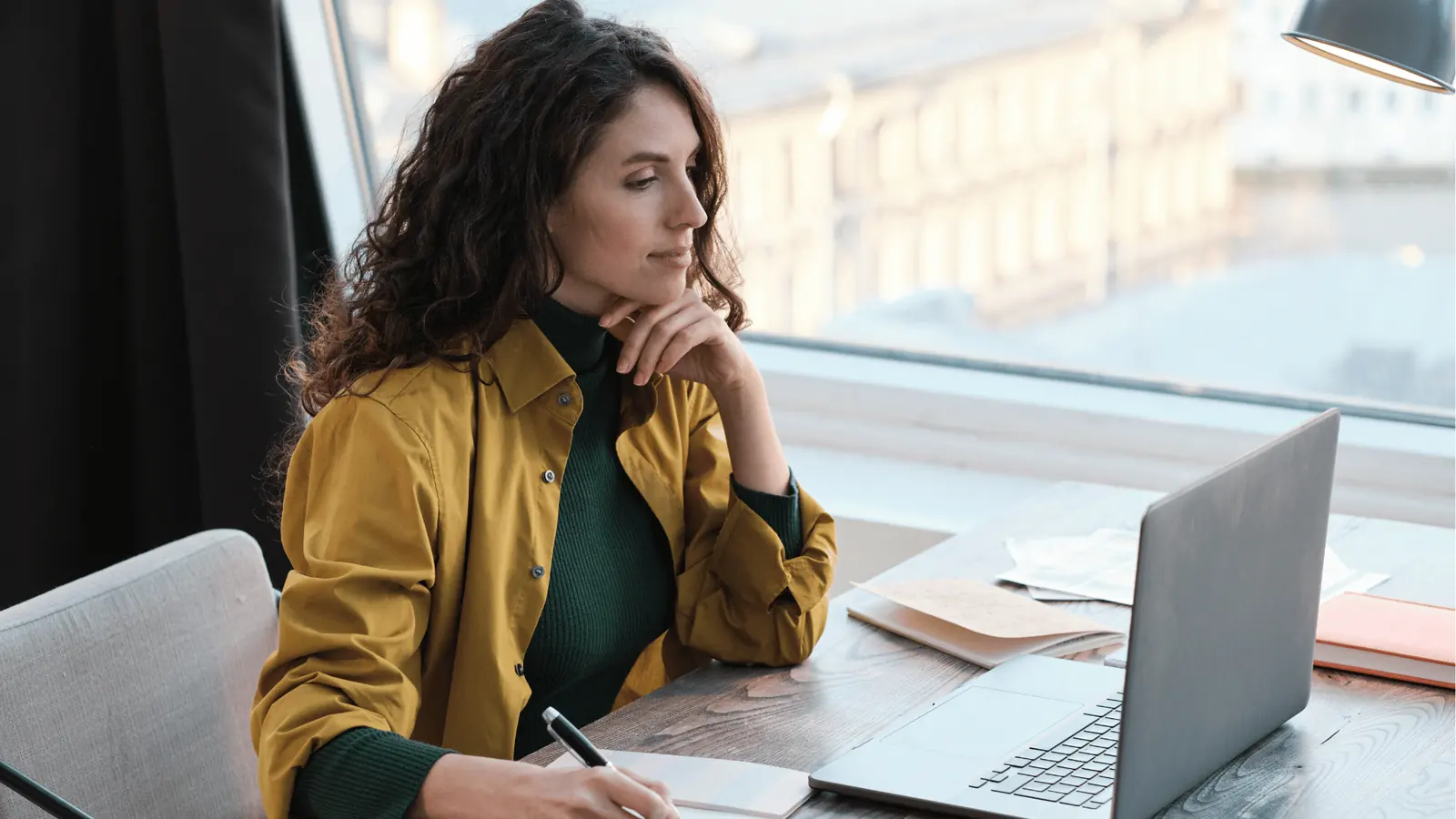 Woman in mustard jacket writing notes while looking at laptop by window