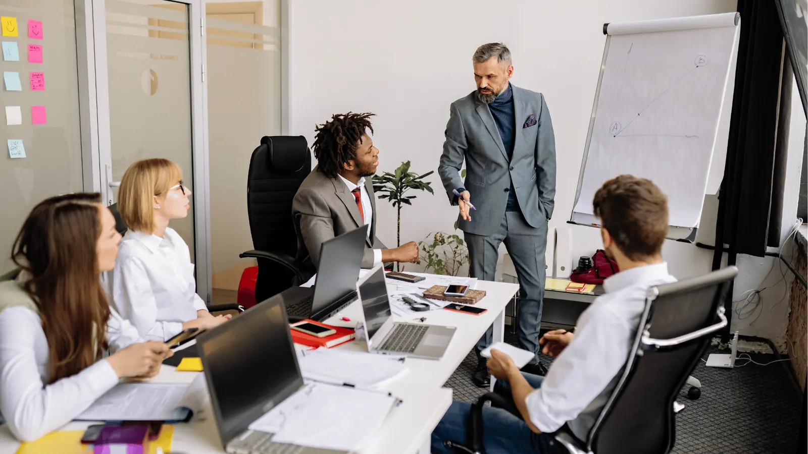 Business team meeting with presenter pointing at colleagues around a laptop-filled table for approval workflow