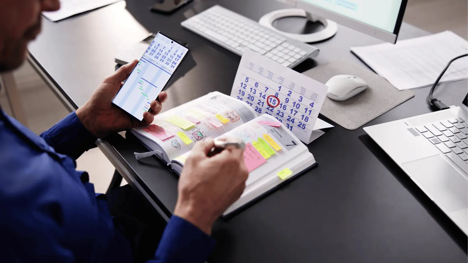 Person writing in a sticky-note filled planner beside phone and desk calendar for approval workflow
