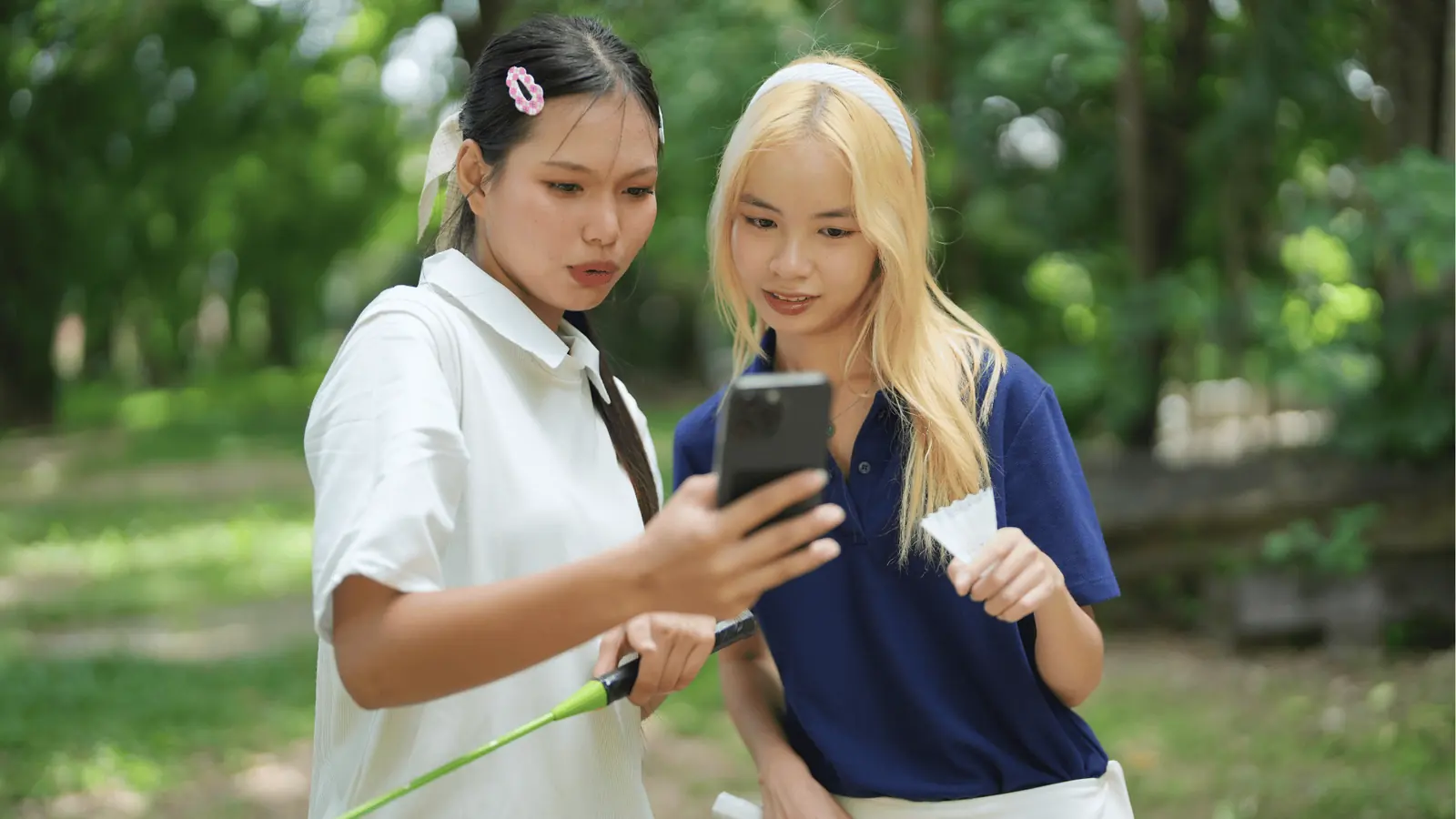 Two girls outdoors looking at a smartphone while holding a shuttlecock