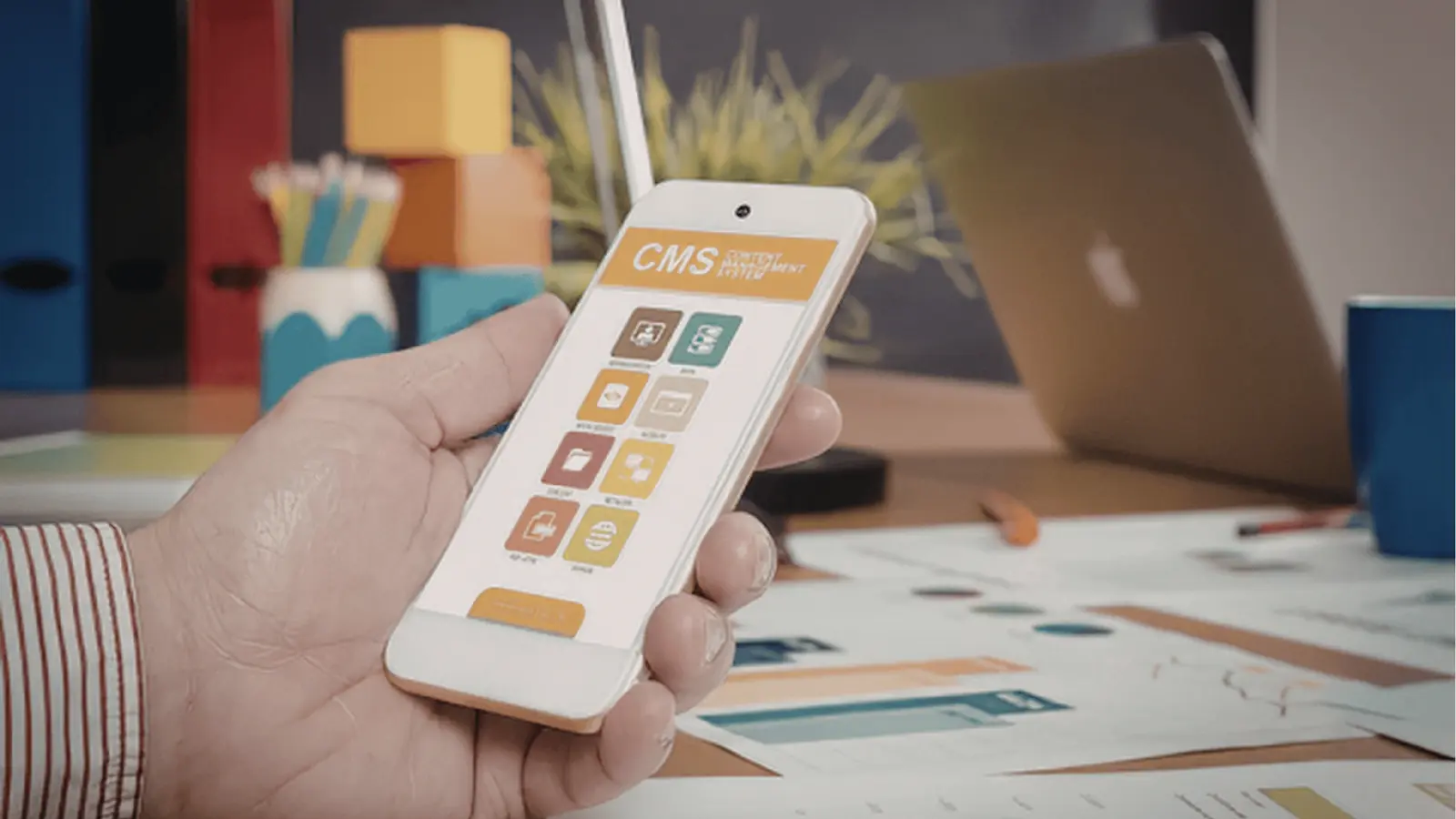 Hand holding smartphone showing CMS app UI over desk with laptop for campaign planning