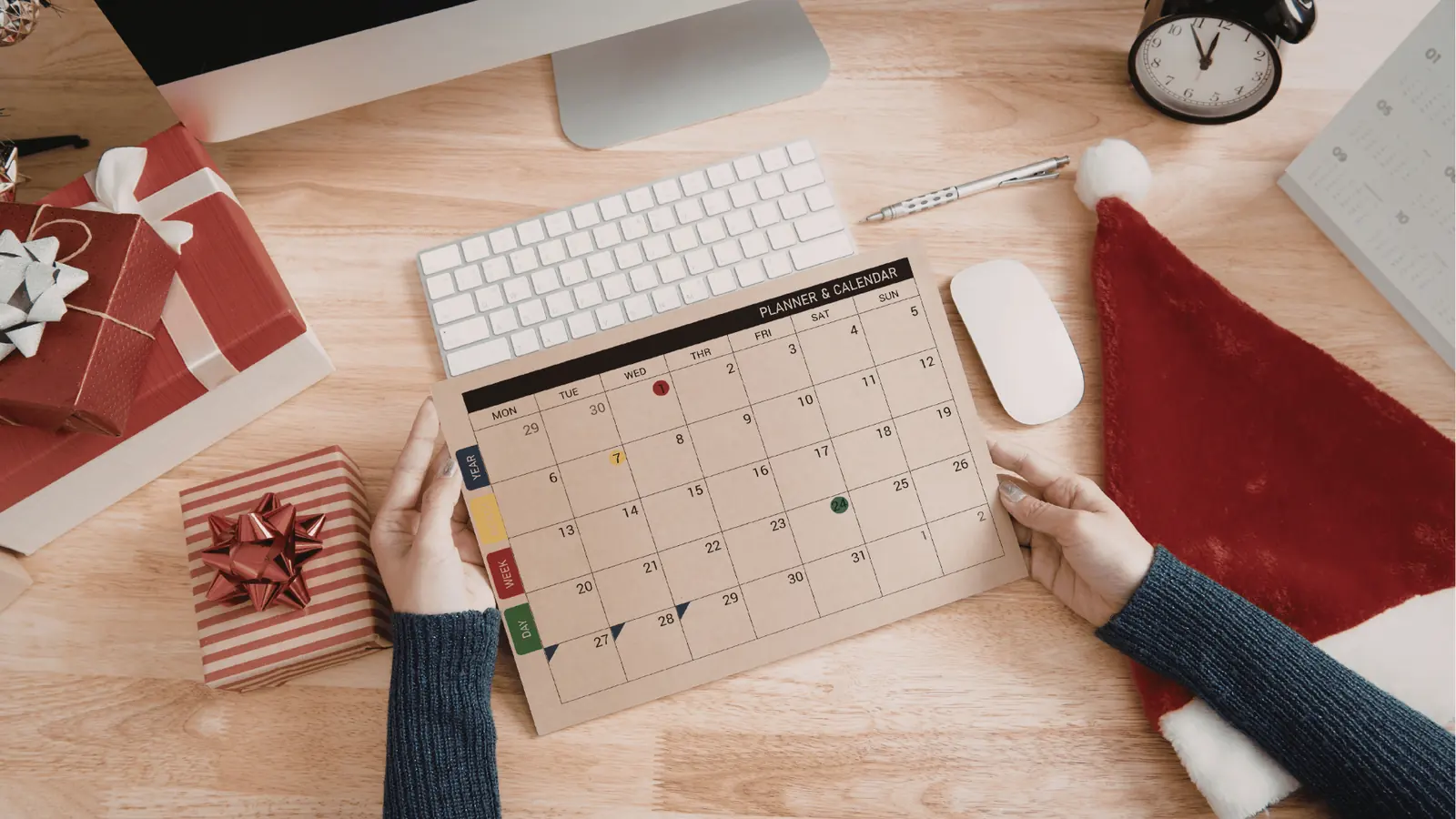 Hands holding a paper monthly planner on a wooden desk with gifts for publishing