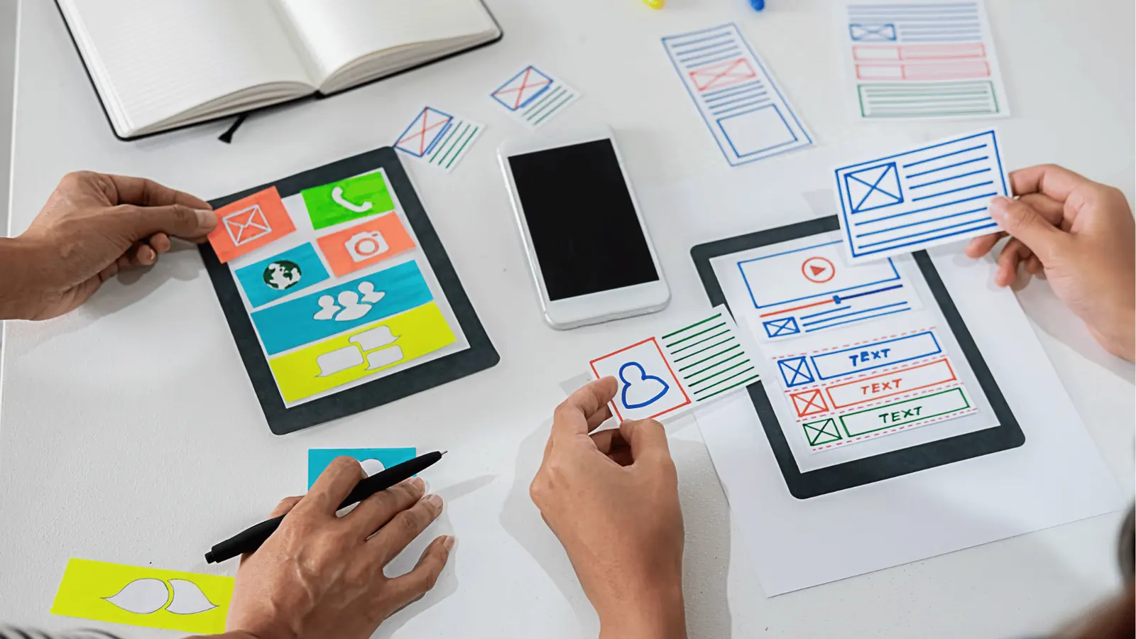 Hands arranging paper mobile app wireframe pieces on a white table