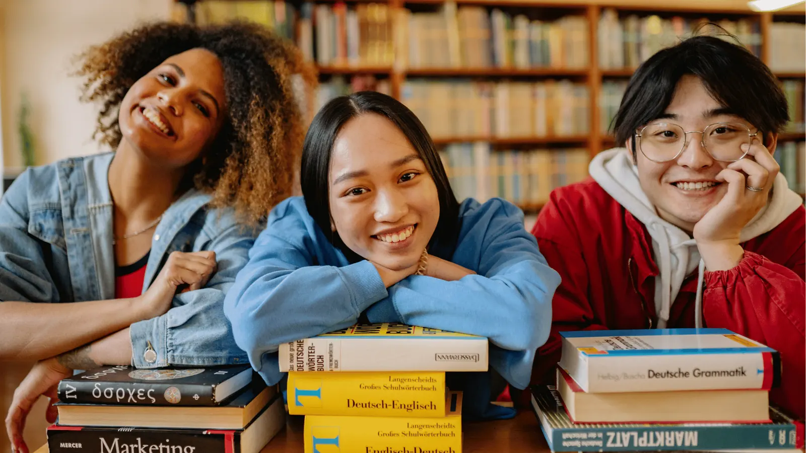 Three smiling students leaning on stacks of books at a library table