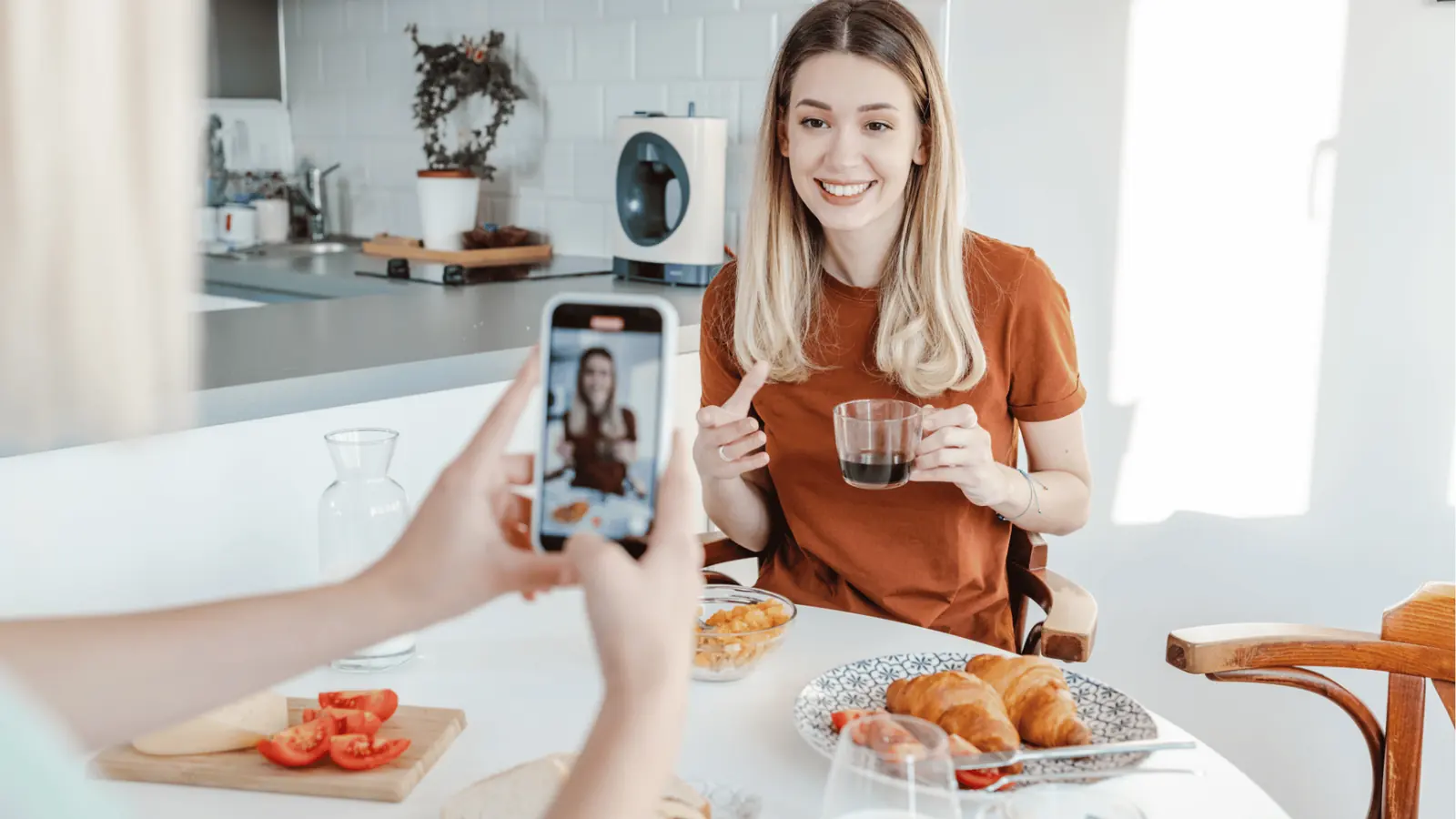 Smiling woman at a breakfast table being photographed on a smartphone while holding coffee