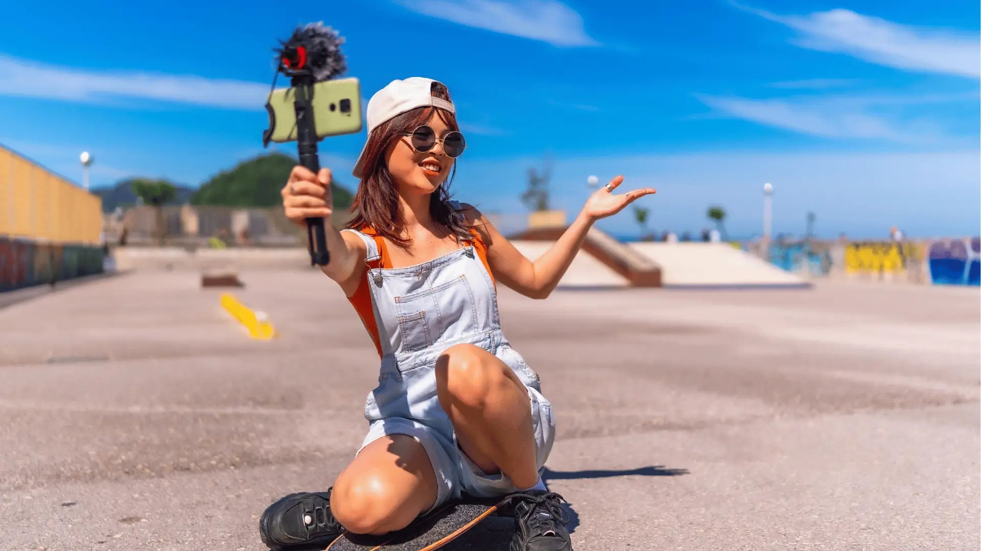 Young woman kneeling on skateboard vlogging with a smartphone mounted on a microphone for approval workflow